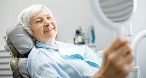 Ältere Frau, liegt auf einem Zahnarztstuhl und betrachtet ihre Prothese.
