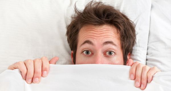 Mann liegt unter einer Bettdecke; nur der obere Teil des Kopfes lugt hervor; die Augen blicken angsterfüllt