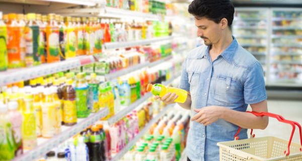 Mann um die 30 im Sommerhemd mit Einkaufskorb am Kühlregal, schaut auf Etikett einer Flasche mit Saft oder Öl