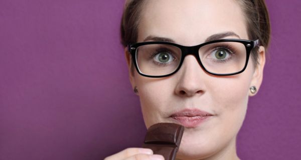 Portraitfoto junge Frau mit großer schwarzrandigen Brille schaut mit aufgerissenen Augen in die Kamera, ein Stück dunkle Schokolade zum Mund führend