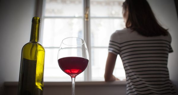 Frau steht am Fenster, hinter ihr stehen eine Weinflasche und Glas mit Rotwein