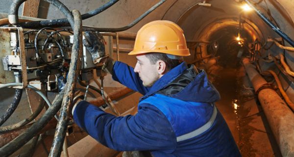 Mann um die 30 in blauer Arbeitskleidung, oranger Schutzhelm, unter Tage in einem Tunnel mit Rohrsystem bei der Arbeit