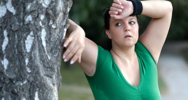 Junge Frau, etwas übergewichtig, im grünen Sportbody, vorne und unter den Armen Schweißflecken, lehnt außer Atem an einem Baum