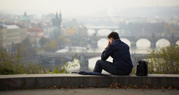 Rückansicht mit Halbprofil eines Mannes, auf einem Mäuerchen sitzend, das Gesicht in die Hände gestützt. Unter ihm die verhangene Stadtansicht von Prag und der Moldau