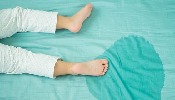 Feuchter Fleck auf einem Bettlaken, daneben Kinderfüße.