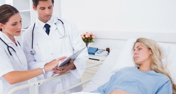 Frau mit langen blonden Haaren im Krankenbett, Ärztin und Arzt mit Clipboard stehen am Bett.