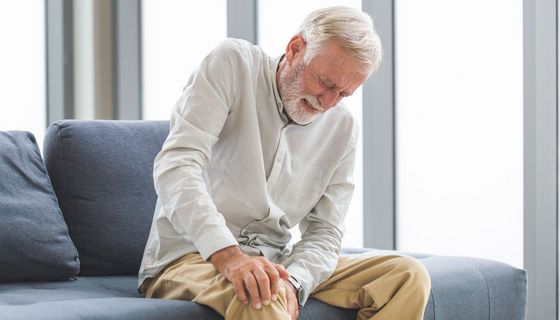Ein älterer Mann sitzt mit schmerzverzerrtem Gesicht auf dem Sofa und hält sich sein Knie.