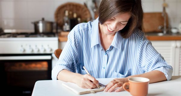 Junge Frau mit brünettem Haar sitzt am Küchentisch und schreibt in ein Journal, lächelnd.