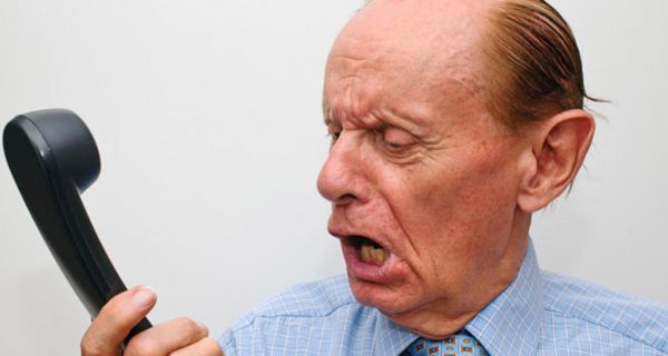 Älterer Mann in blauem Hemd und Schlips schaut wütend auf einen Telefonhörer in seiner Hand und brüllt hinein