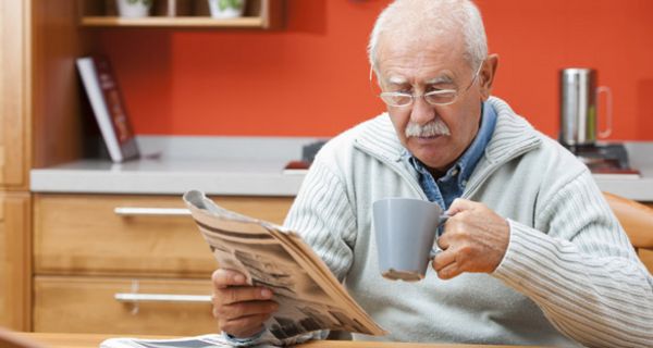 Senior am Küchentisch beim Zeitunglesen, in der rechten Hand die Zeitung, in der linken einen Kaffeehumpen