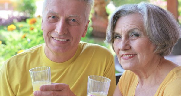Sommerszene: Paar in den 60ern, Portrait, in die Kamera schauend, gelbe T-Shirts, jeweils ein Glas Limo in der Hand. Beide lächeln