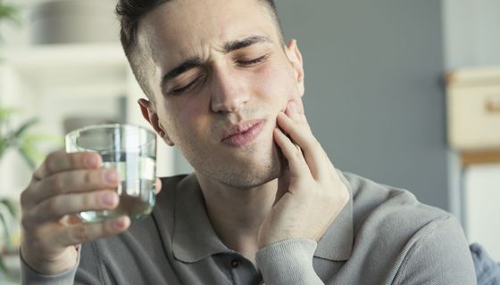 Ein junger Mann hält sich mit schmerzverzerrtem Gesicht die Wange, in der anderen Hand ein Wasserglas.