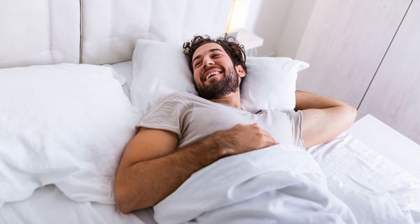 Junger Mann mit braunen Locken liegt im Bett und lächelt, ist ausgeschlafen.