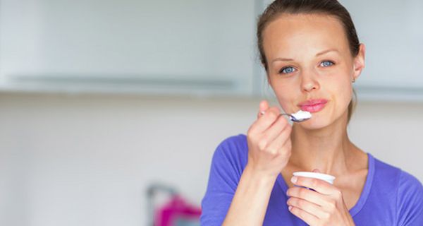 Frontalfoto Frau ca. 30 in lilablauem Shirt, blaue Augen, rosa geschminkte Lippen, Haare zurückgebunden, löffelt genussvoll einen Joghurt