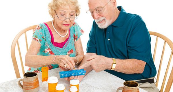 Farbenfroh gekleidetes Seniorenpaar am Tisch bei der Einteilung der Wochenration Tabletten in eine Dosierhilfe