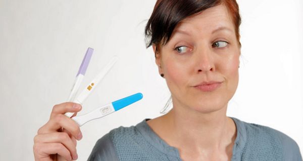 Jüngere Frau mit zusammengebundenen, braunen Haaren, grauer Pulli, schaut skeptisch auf 3 Schwangerschaftstests in ihrer rechten Hand
