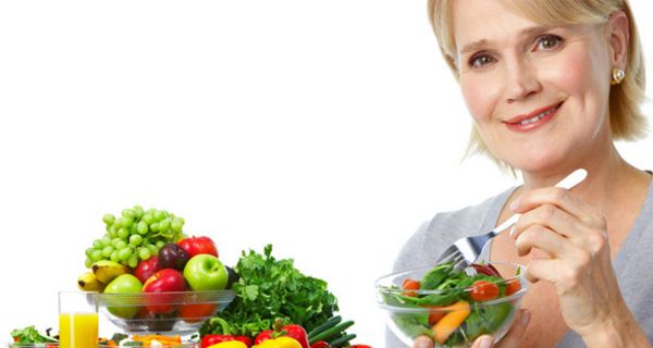 Blonde Frau um die 50 isst ein Schälchen mit gemischtem Salat; auf dem Tisch Obstschale und darum herum verschiedene Gemüse, eine Müslischale und ein Glas Orangensaft
