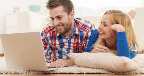 Junges, attraktives Paar, auf weißem Teppich liegend, schaut vergnügt in einen Laptop