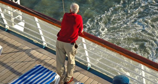 Rückenansicht eines älteren Mannes im roten Pulli und heller Hose an der Reeling eines Kreuzfahrtschiffes lehnend und aufs Meer schauend