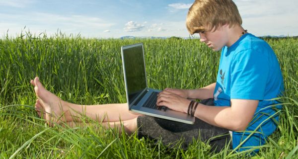 Blonder Teenagerjunge, ca. 15,  in blauem Shirt auf grüner Wiese mit einem Laptop auf den Beinen