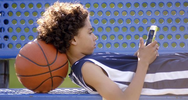 Junge, liegt mit Smartphone auf einer Bank und stützt seinen Kopf auf einen Basketball.