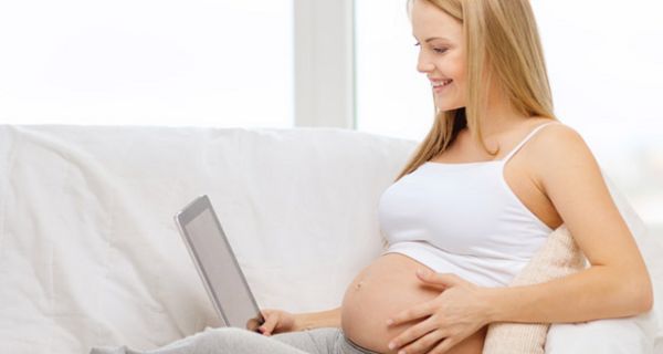 Schwangere Frau in grauer Gym-Hose und bauchfreiem Oberteil, blonde Haare, auf weißer Couch mit Laptop, eine Hand fasst an den Bauch