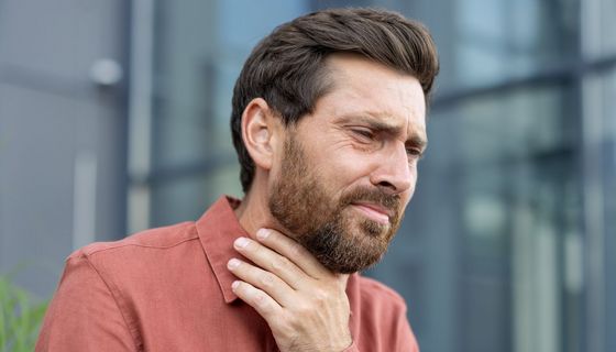 Mann gegen Ende 30 legt sich die Hand an den Kehlkopf, er sieht nicht glücklich aus.