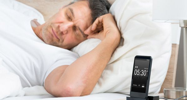Mann um die 40 schläft auf der Seite im weißbezogenen Bett, weißes T-Shirt, Vordergrund Nachttisch mit Smartphone in Ladestation