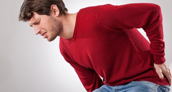 Dunkelhaariger Mann um die 30, Jeans, dunkelroter Pullover, hält sich mit schmerzverzerrtem Gesicht den unteren Rücken