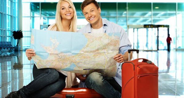 Frau und Mann, lachend, ca. 30+, Sitzen auf rotem Koffer in Flughafenabflughalle, roter Koffer neben Mann, und halten Landkarte in den Händen