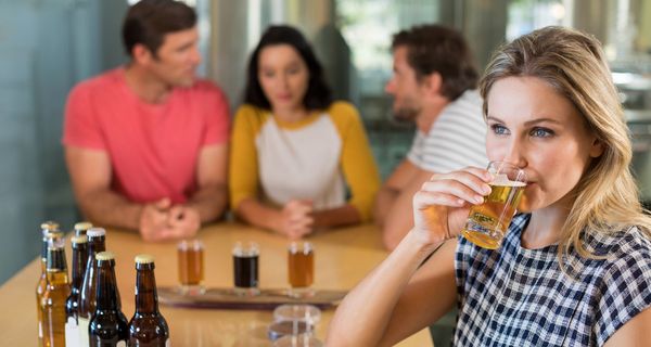 Junge Frau trinkt ein Glas eines alkoholischen Getränks und schaut dabei nachdenklich in die Ferne. Ihre Freunde im Hintergrund unterhalten sich. 