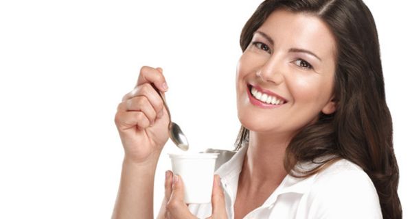 Frau mit dunkelen, langen Haaren, lächelt in die Kamera, während sie Joghurt löffelt