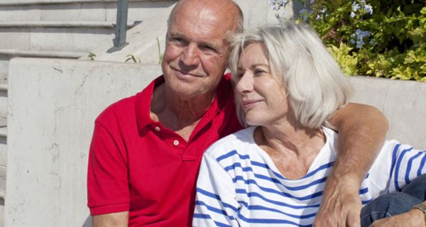 Älteres Paar sitzt in der Sonne; er im roten Polo-Shirt hat den Arm um ihre Schulter gelegt; sie trägt ein blau-weiß gestreiftes Top
