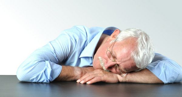 Älterer Mann im hellblauen Hemd schläft am Tisch mit dem Kopf auf den Armen.