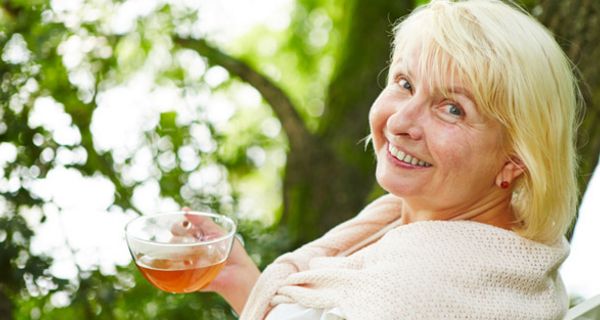 Ältere Frau sitzt in ihrem Garten und hält eine Tasse Tee in der Hand.