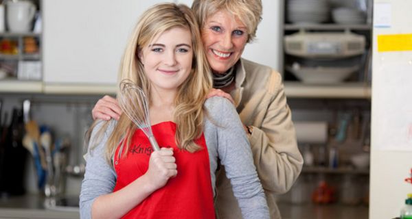 Küchenszene: Teenagertochter im Vordergrund, blonde Haare, rote Schürze, Schneebesen in der rechten Hand, dahinter sie an den Schultern haltend Mutter, beide in die Kamera lächelnd