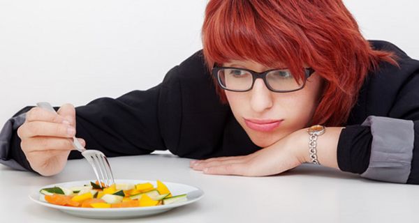 Junge rothaarige Frau, schwarzrandige Brille, guckt missmutig über Tischkante auf Teller mit geschnittenem Obst