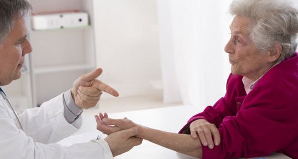 Arzt (linker Bildrand) erklärt Seniorin (rechts) etwas, dabei betrachtet und hält ihre Hand