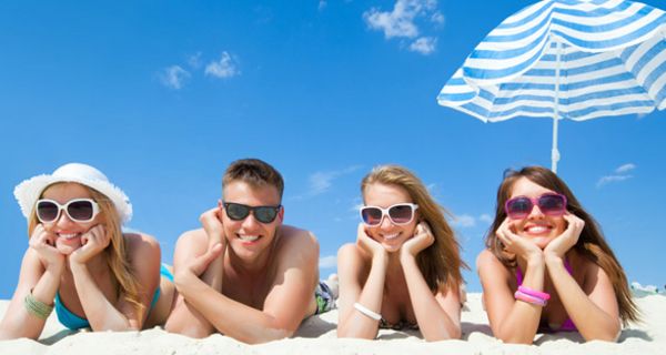 Vier junge Erwachsene liegen am Strand auf dem Bauch und tragen Sonnenbrillen, im Hintergrund ein Sonnenschirm