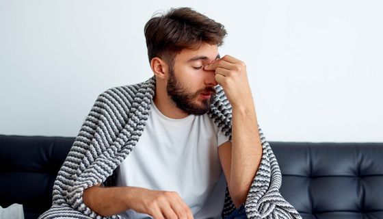 Mann mit Nasennebenhöhlenentzündung sitzt mit Decke und Taschentuchspender auf einer Couch. Er hält sich den Nasenrücken.