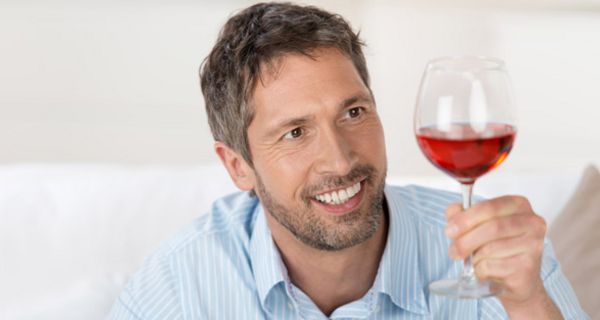 Mann mit Dreitagebart im hellblau-weiß gestreiften Hemd betrachtet lächelnd ein bauchiges Glas, gefüllt mit Rotwein
