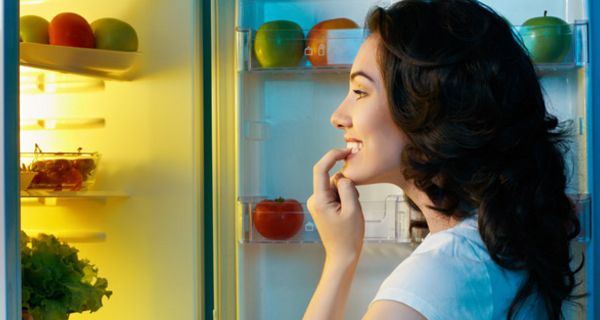 Frau um die 30 mit dunklen Haaren vor dem geöffneten Kühlschrank beißt sich auf einen Fingrnagel nach dem Motto: soll ich oder soll ich nicht?