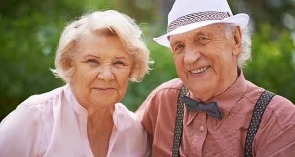 Portraitbild: freundlich lächelndes Seniorenpaar, schicke Freizeitkleidung (Mann Fliege, rotkariertes Hemd, weißer Strohhut, Hosenträger, Frau gut frisiert, hellrosa kragenlose Bluse)