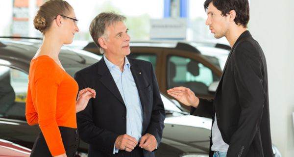 Junges Paar in Verhandlung mit einem älteren Autoverkäufer, Autos im Hintergrund