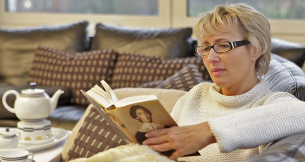 Frau in den mittleren Jahren mit Kurzhaarschnitt und Brille sitzt auf einer Couch und liest ein Buch
