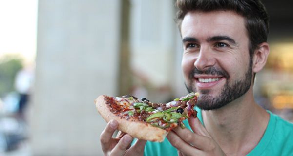 Foto Gesicht und Schulterbereich, junger dunkelhaariger, bärtiger Mann hält sich lächelnd ein großes Stück Pizza vor den Mund.