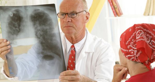 Frau mit rotem Pullover und rotweiß gemustertem Kopftuch Halbprofil, Arzt von vorne, der ernst auf ein Röntgenbild schaut, das den Brustkorb zeigt
