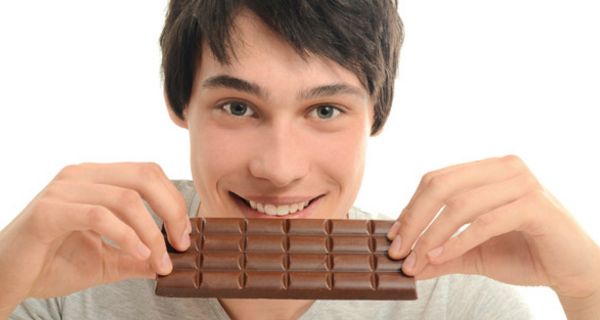 Frontalfoto Portrait junger Mann im grauen T-Shirt, der mit beiden Händen eine Tafel Schokolade quer zum Mund führt, um reinzubeißen