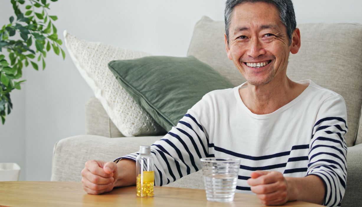 Älterer Mann mit einer Packung Tabletten und einem Glas Wasser, lächelt in die Kamera.