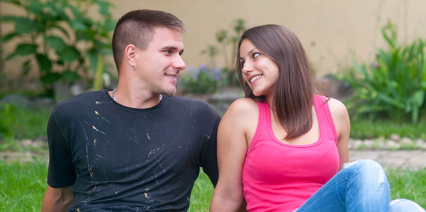 Junges Paar auf einer Wiese nebeneinander sitzend (Jeans, Shirts) schaut sich verliebt in die Augen